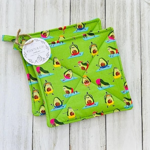 May include: Two green pot holders with a pattern of avocados doing yoga. The pot holders are folded and have a white tag with the text "Mountain Girl Creations".