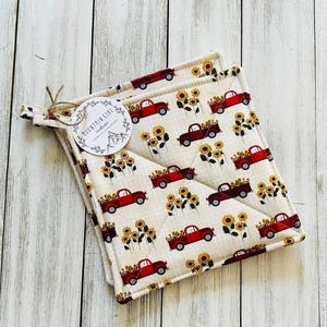 May include: Two square pot holders with a repeating pattern of red trucks carrying sunflowers on a cream background. The pot holders are quilted and have a loop for hanging. A tag with the text "MOUNTAIN GIRL traditions" is attached.