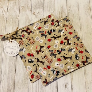 May include: Two beige pot holders with a pattern of dogs and bones. The pot holders have red trim and a white tag with the text "Mountain Girl Creations".