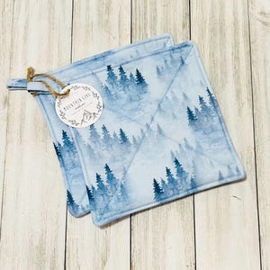 May include: Two square, light blue pot holders with a forest scene design. The pot holders have a quilted pattern and a loop for hanging. A round tag with the text "Mountain Girl Creations" is attached.