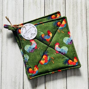 May include: Two green square pot holders with a rooster pattern. The roosters are red, blue, yellow, and orange. The pot holders are tied together with twine and a tag that says "Mountain Girl creations".