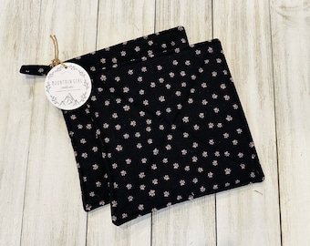 May include: Two black quilted pot holders with a pink paw print pattern. The pot holders have a loop for hanging. The tag attached to the pot holders reads "Mountain Girl Creations".