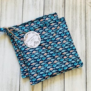 May include: Two navy blue pot holders with a fish pattern in teal and coral. A tag with the text "MOUNTAIN GIRL creations" is attached with twine. The pot holders are square and have a loop for hanging.