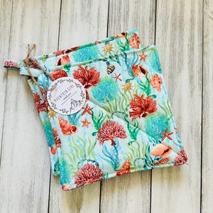 May include: Two square pot holders with a light blue background and a colorful sea life pattern. The design includes red coral, starfish, seashells, and seaweed. A tag with the text "MOUNTAIN GIRL creations" is attached with twine.