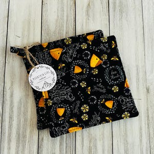 May include: Two black pot holders with a bee and honeycomb pattern. The pot holders have white text that says "Bee Yourself" and "Bee Kind".
