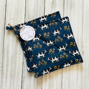 May include: Two square pot holders with a dark teal background featuring a pattern of black and white cows and yellow sunflowers. The pot holders have a loop for hanging and a tag that reads "MOUNTAIN GIRL creations".