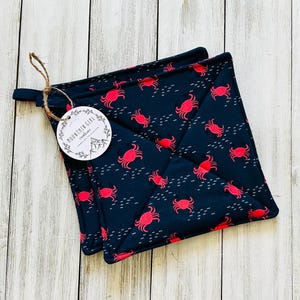 May include: Two navy blue square pot holders with a red crab pattern and white accents. A tag with the text "MOUNTAIN GIRL creations" is attached with twine. The pot holders are on a light wood surface.