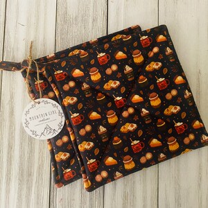 May include: Two black pot holders with a fall-themed print of mugs, pies, and cinnamon sticks. The pot holders have a white tag with the text "Mountain Girl Creations".