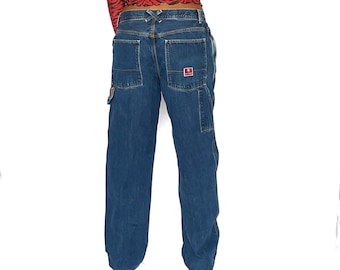 May include: A pair of blue denim carpenter jeans with a red logo patch on the back pocket. The jeans have a loose fit and are worn by a person with a red and black striped shirt.