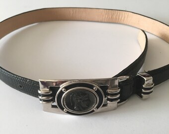 May include: A black leather belt with a silver buckle featuring a round, black, raised design of a warrior's head.