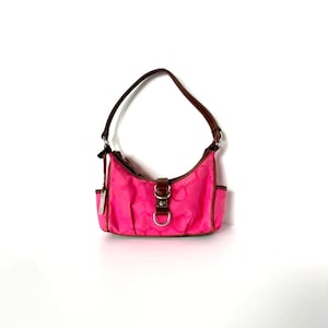 May include: A small hot pink handbag featuring a brown leather-like strap and trim. The bag has a central brown tab with a silver-tone metal buckle and D-ring closure. The exterior fabric displays a subtle repeating pattern and side pockets.