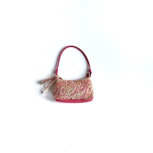 Puede incluir: Un pequeño bolso rosa con un estampado de cachemira en tonos rosa y beige. El bolso tiene un asa y una base rosas a juego. Un lazo decorativo con detalles plateados adorna el asa.