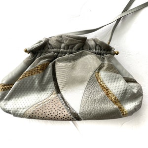 Puede incluir: Un bolso de mano de cuero gris con cierre de cordón y una correa larga. El bolso está hecho de diferentes tipos de cuero, incluyendo piel de serpiente y cuero perforado.