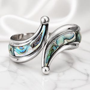 Può includere: Un bracciale a polsino argentato con un design aperto. Il bracciale presenta sezioni curve intarsiate con conchiglia di abalone iridescente. Ogni estremità del bracciale ha una piccola sfera argentata rotonda.