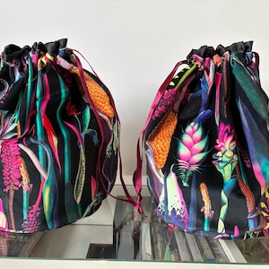 Op de afbeelding: Twee trekkoordtassen met een levendig bloemenpatroon op een zwarte achtergrond. De tassen hebben een kleurrijk ontwerp met roze, oranje, groene en blauwe botanische elementen. Elke tas heeft een zwarte bovenkant en een bordeauxrood trekkoord.