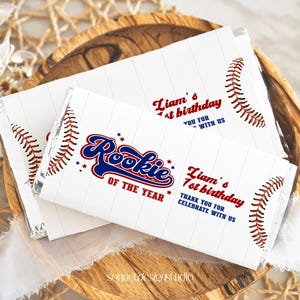 May include: White chocolate bar wrapper with a baseball theme. It says "Liam's 1st birthday" and "Rookie of the Year" with red, white and blue baseball stitching.