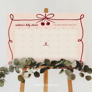 May include: A light pink baby shower calendar with a dark red cherry design and text. The calendar has a grid for tracking dates and a decorative border. A wooden easel supports the calendar, with green foliage at the base.