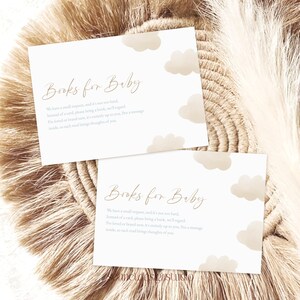 May include: Two cream-coloured baby shower cards with the text "Books for Baby" and cloud illustrations. The cards have a beige background and are placed on a woven, beige surface.