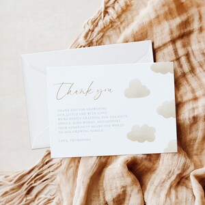Może przedstawiać: Biała kartka z podziękowaniami z pasującą kopertą. Karta zawiera słowa "Thank you" w eleganckim skrypcie, wraz z przesłaniem wdzięczności i ilustracjami chmur. Karta jest umieszczona na tle beżowej tkaniny.