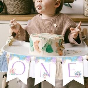 May include: A white and purple banner with the word "ONE" in purple letters. Each letter has a mermaid or dolphin illustration. The banner is decorated with blue and gold glitter and has white tassels.