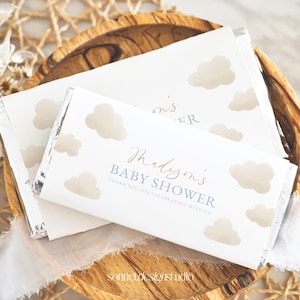 May include: White chocolate bar wrappers with a cloud design and the text "Madison's Baby Shower". The wrappers are silver on the inside and are arranged on a wooden bowl. The design includes light brown clouds.
