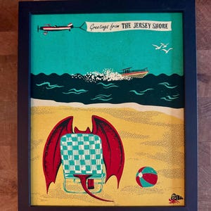 May include: A framed print featuring a beach scene with a winged creature sitting in a checkered beach chair on yellow sand. A boat travels on dark water, and an airplane pulls a banner that reads Greetings from THE JERSEY SHORE.