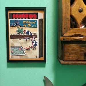May include: Framed artwork depicting a seaside scene with a pool, beach umbrellas, and the text "Wildwood by the sea." The artwork is in a black frame and hangs on a teal wall. A wooden cabinet is to the right.