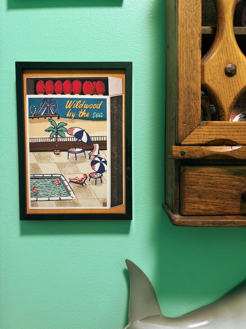 May include: Framed artwork depicting a seaside scene with a pool, beach umbrellas, and the text "Wildwood by the sea." The artwork is in a black frame and hangs on a teal wall. A wooden cabinet is to the right.