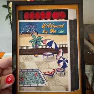 May include: Framed artwork featuring a beach scene with a Ferris wheel, palm tree, and pool. The text "Wildwood by the sea" is displayed in blue and yellow. The artwork is in a black frame.
