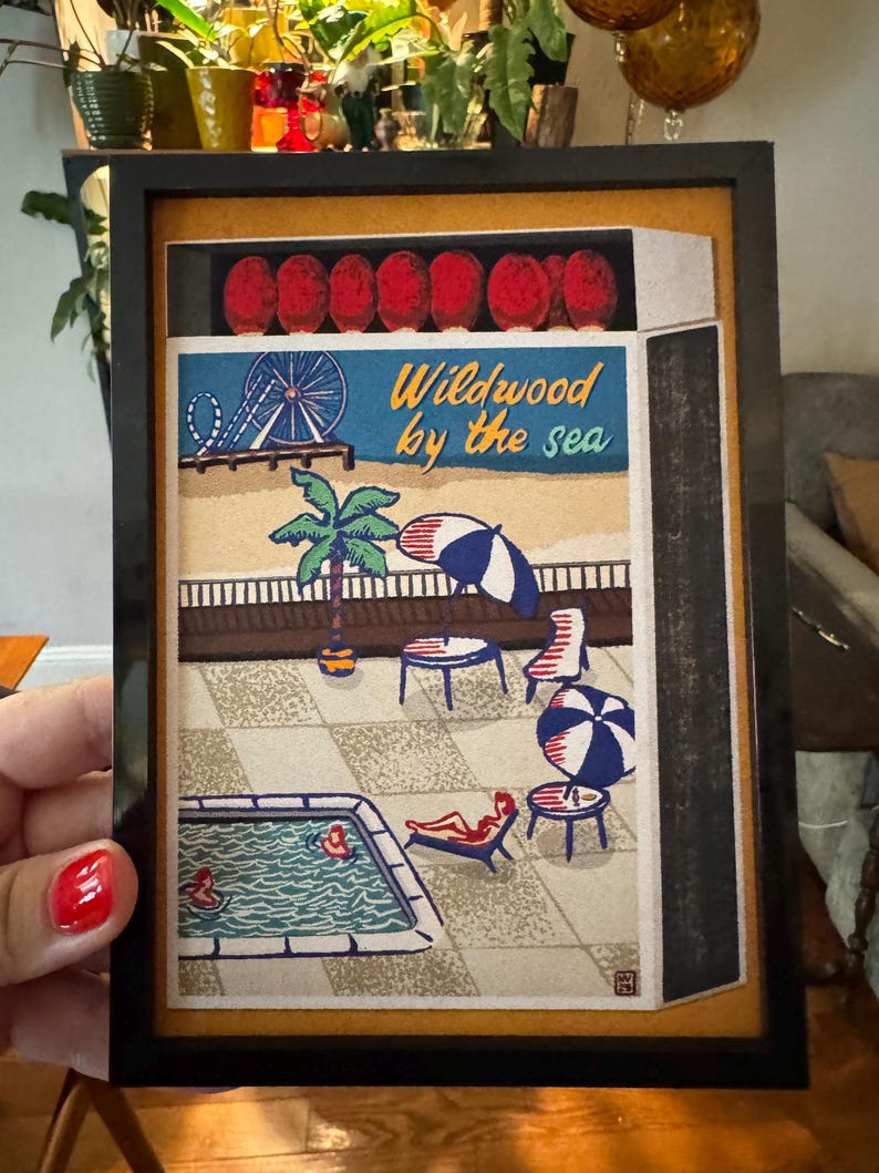 May include: Framed artwork featuring a beach scene with a Ferris wheel, palm tree, and pool. The text "Wildwood by the sea" is displayed in blue and yellow. The artwork is in a black frame.