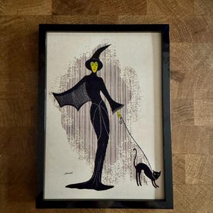 May include: Framed art print featuring a stylized illustration of a witch in a long black dress and hat, holding a leash attached to a black cat. The witch has a yellow face and is set against a patterned background.