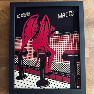 May include: Framed art print featuring a red devil-like figure seated at a diner counter. The artwork has a black background with the words "ICE CREAM" and "MALTS" in white. The counter is red and white, with black and white checkered flooring.