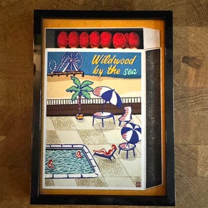 May include: A framed artwork depicting a seaside scene with a pool, beach umbrellas, and the text "Wildwood by the sea." The artwork features a Ferris wheel, palm trees, and people relaxing by the pool. The frame is black.