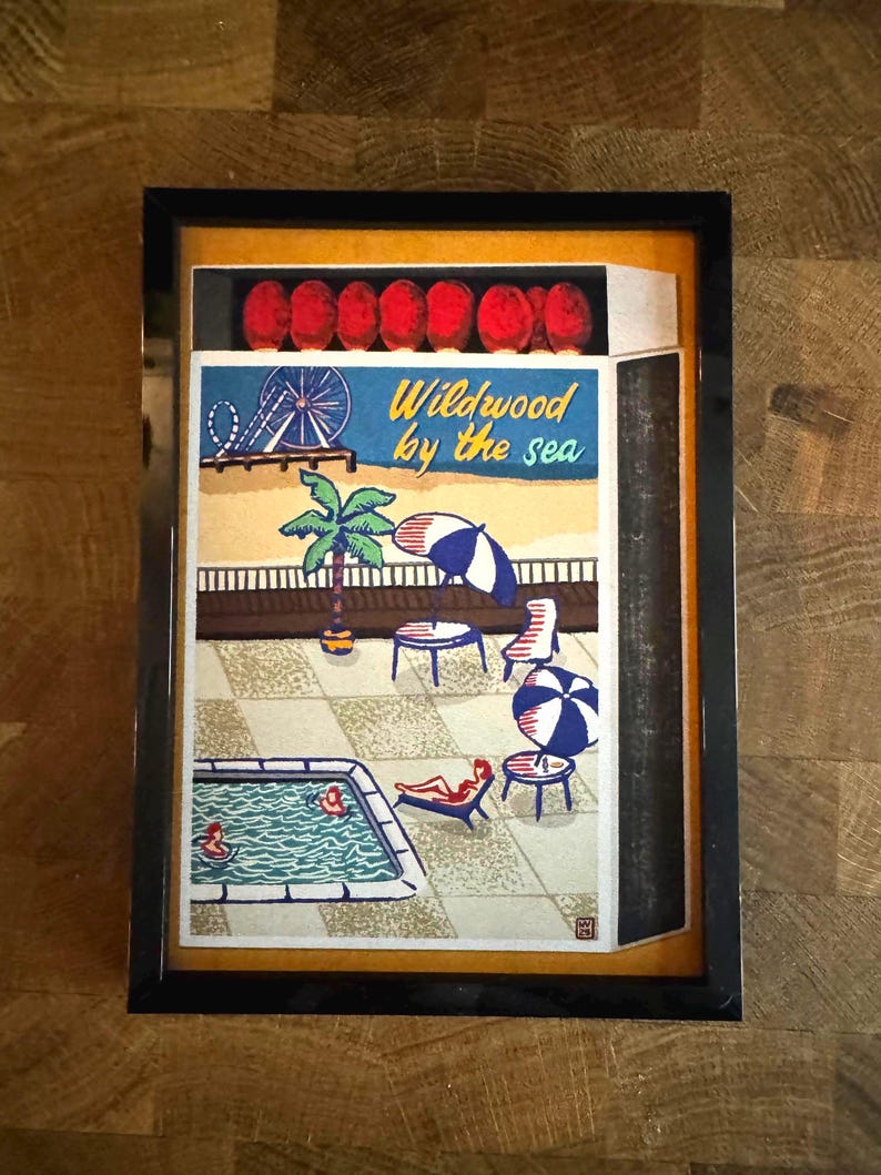 May include: A framed artwork depicting a seaside scene with a pool, beach umbrellas, and the text "Wildwood by the sea." The artwork features a Ferris wheel, palm trees, and people relaxing by the pool. The frame is black.