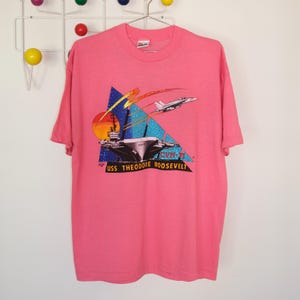 May include: Pink t-shirt featuring a graphic of the USS Theodore Roosevelt aircraft carrier with a jet flying overhead. The design includes a sunset, geometric shapes, and the text "CVN-71".