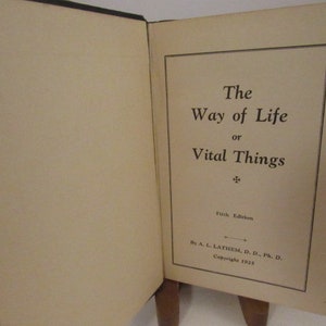 May include: An open book with the title "The Way of Life or Vital Things" printed on the right page. The book is a fifth edition, copyrighted in 1925. The pages are off-white, and the cover is black.