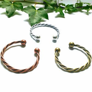 May include: Three open cuff bracelets in silver, copper, and brass tones. Each bracelet features a twisted rope design and a small ball at each open end. The bracelets are displayed on a white surface with green foliage in the background.