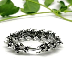 Könnte beinhalten: Silberfarbenes Armband mit einem wirbelsäulenartigen Design. Das Armband besteht aus einer Reihe von miteinander verbundenen, stilisierten Wirbeln, die ein einzigartiges Accessoire bilden. Das Metall hat ein leicht gealtertes Aussehen, das die komplizierten Details hervorhebt.