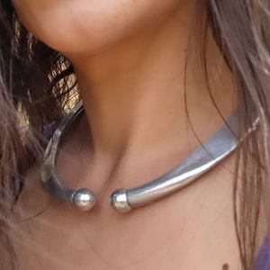 Könnte beinhalten: Eine silberne Choker-Kette mit abgerundeten Enden. Die Kette besteht aus einem dicken, flachen Metallband.