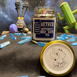 May include: A glass jar filled with tea, labeled "Aether Butterfly Rose White Tea." The jar sits on a black surface with blue crystal-like objects scattered around. A gold-rimmed lid with an elephant logo is in the foreground. The background features a space-themed backdrop.