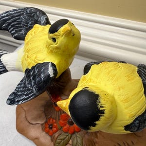 Puede incluir: Una figura de cerámica con dos pájaros amarillos y negros posados sobre una base de rama marrón con flores naranjas y hojas verdes. Los pájaros tienen cuerpos de color amarillo brillante, cabezas negras y alas negras con detalles blancos, vista cenital.