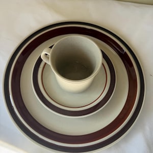 May include: A beige ceramic cup and saucer set with brown and black striped accents. The set includes a large plate, a smaller plate, and a cup. The plates and cup have a speckled texture.