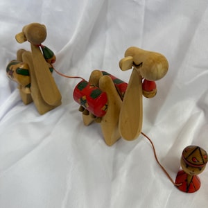 May include: Wooden camel toy set with a pull-along design. The camels are light brown with red and green accents on their backs. A small wooden figure in red and brown is attached by a string, completing the set. The toy is set against a white background.