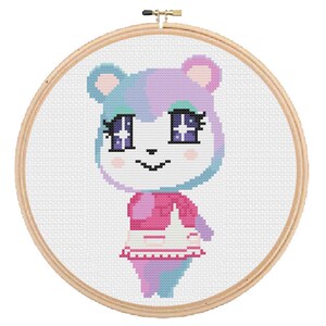 May include: Cross-stitch embroidery hoop featuring a cartoon character with pink and blue fur, wearing a pink and white dress. The character has large eyes with purple and white stars.