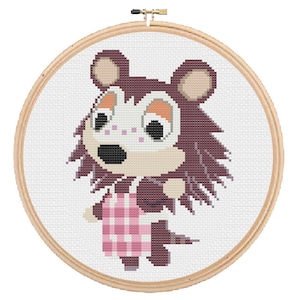 May include: Cross-stitch embroidery hoop featuring a cartoon animal character with brown fur, a pink and white checkered apron, and large eyes. The character is a wolf or a dog.