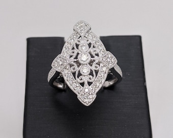 May include: A silver ring with a diamond-shaped design set with small diamonds. The ring is displayed on a black ring display stand.