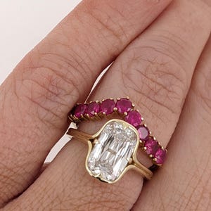 May include: A gold ring set featuring a central emerald-cut clear stone in a bezel setting paired with a curved band adorned with a row of round, vibrant pinkish-red stones. The two rings are worn together, creating a nested, layered appearance.