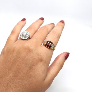 May include: Two gold rings with gemstones. The ring on the left has a large, round, clear stone surrounded by smaller clear stones. The ring on the right has a red stone surrounded by smaller clear stones.