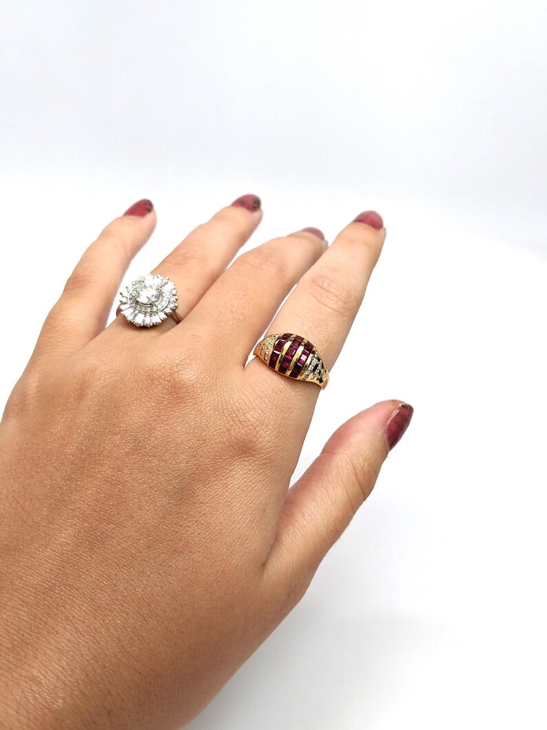 May include: Two gold rings with gemstones. The ring on the left has a large, round, clear stone surrounded by smaller clear stones. The ring on the right has a red stone surrounded by smaller clear stones.
