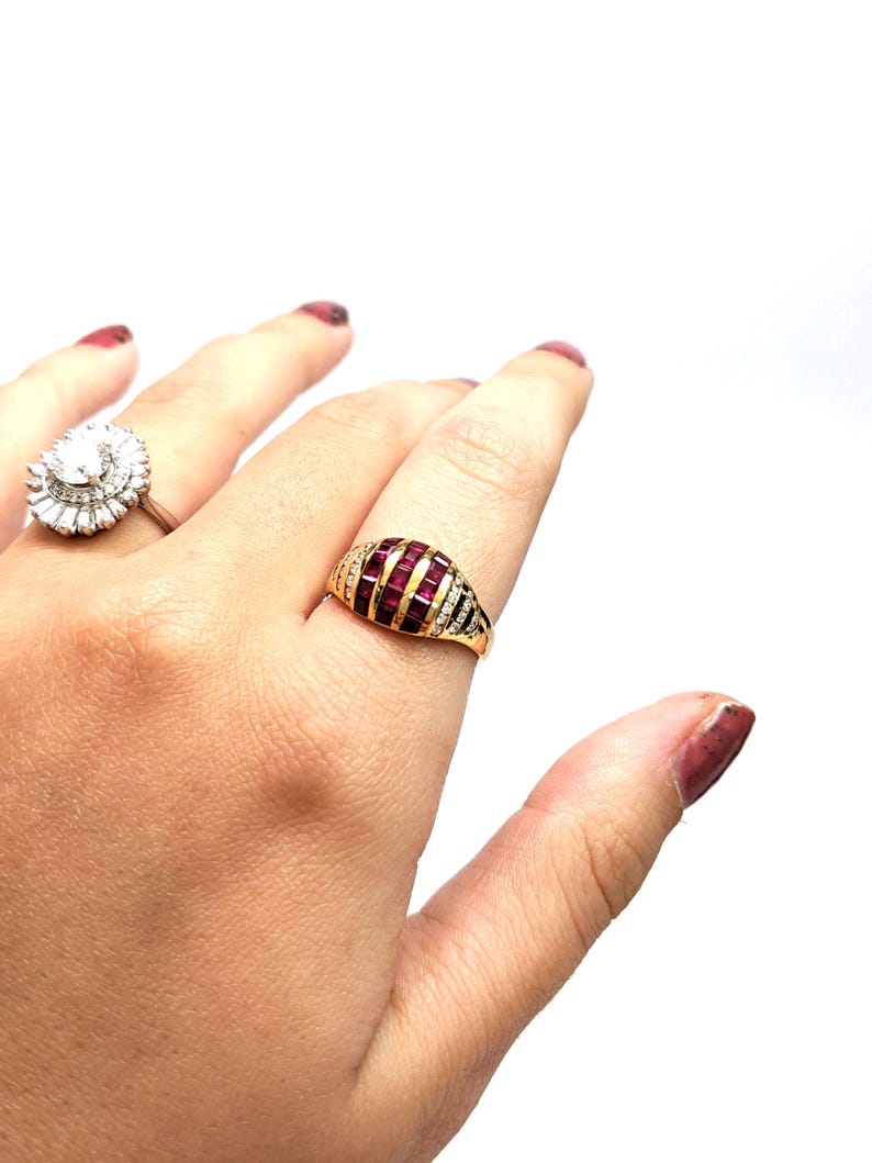 May include: A gold ring with a red stone set in a dome shape. The stone is surrounded by smaller white stones.